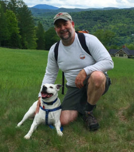 Rick kneeling while hiking with woodlands in background and Leo the white pitbull laying down in front.